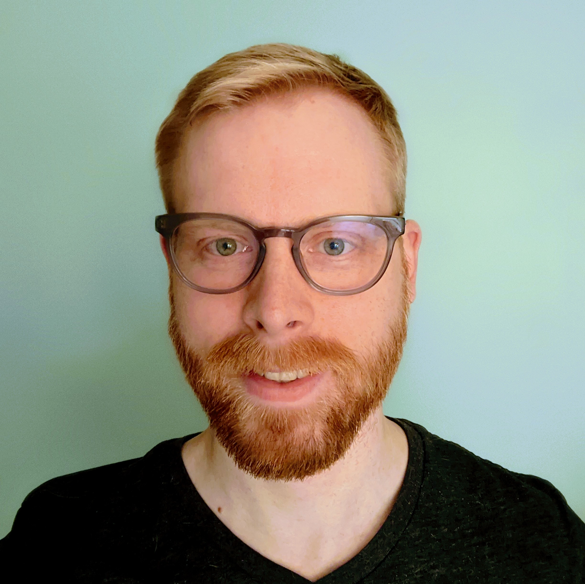 A portrait of a light skinned man with a short red beard and blond hair. He is wearing glasses and a black v-neck tee shirt.